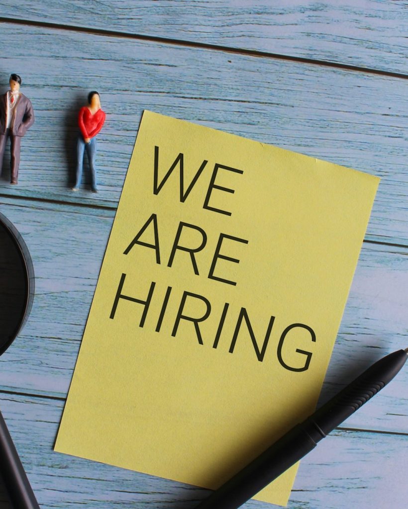 Miniature people and magnifying glass with text WE ARE HIRING